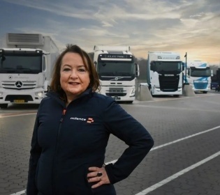 Anja van Niersen smiles into the camera, 5 stationary trucks can be seen in the...