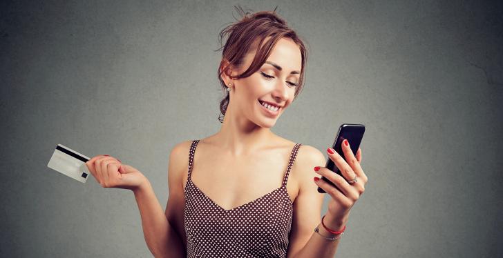 Woman in a dress is smiling at her smartphone and has a card in her hand;...