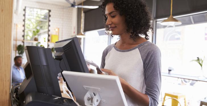 Dark-skinned woman in white-grey top in front of a cash register with several...