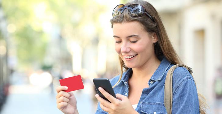 young woman looking at her smartphone while holding a gift card in her hand...