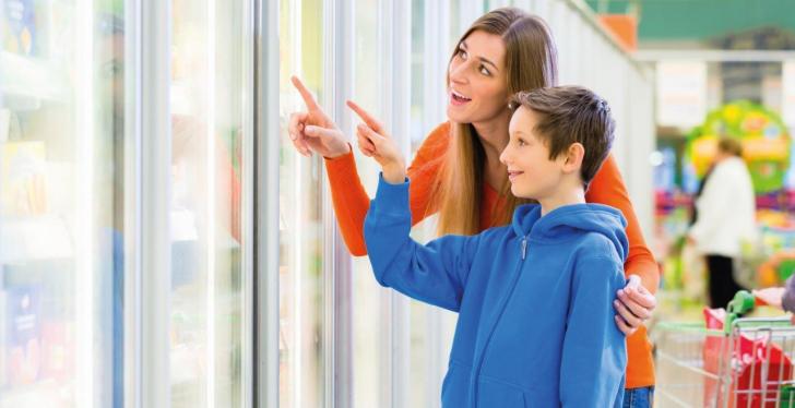 A mother and a boy point their fingers at a refrigerated shelf in a supermarket...