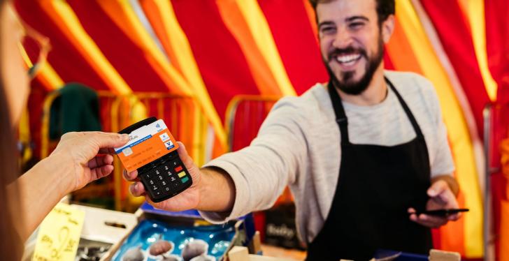 A fruit and vegetable retailer at a market stand holds out a mobile payment...