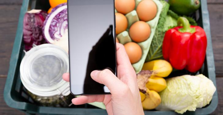 A hand holds a smartphone and behind it is a basket of fresh food...