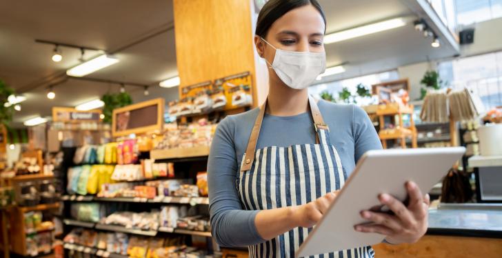 A saleswoman with an apron and face mask holds a Tablet PC in her hand...