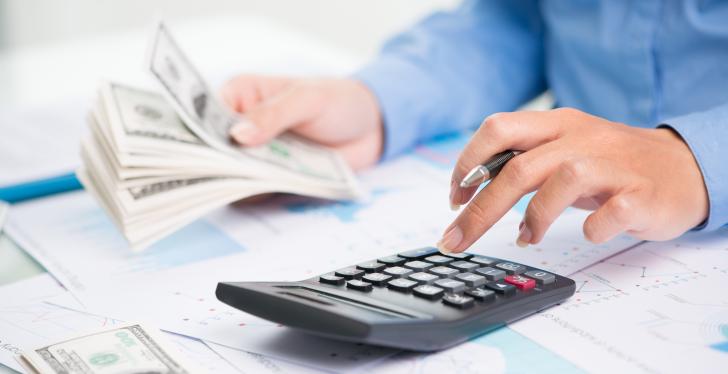 Man is counting the stack of paper money in his left hand with a calculator in...