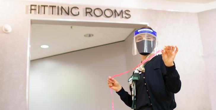 A woman with face mask and measuring tape in front of a fitting room...