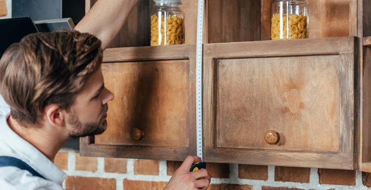 A young man measures the height of a wooden hanging shelf with a tape measure;...