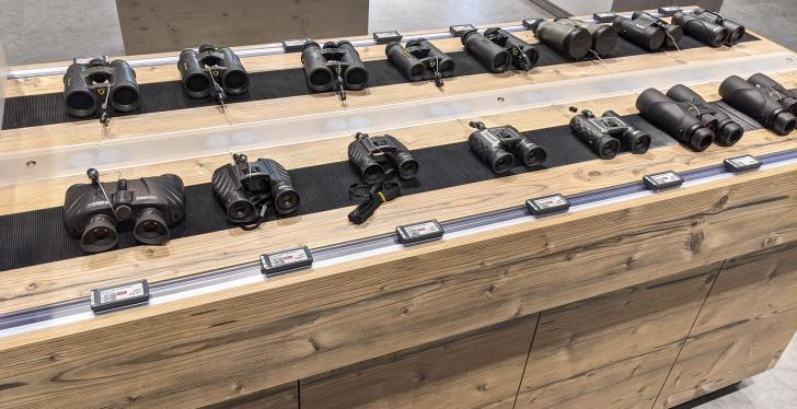 Many binoculars lie side by side on a counter with electronic price tags...