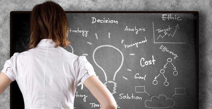 A smartly dressed woman stands in front of a fully written blackboard...