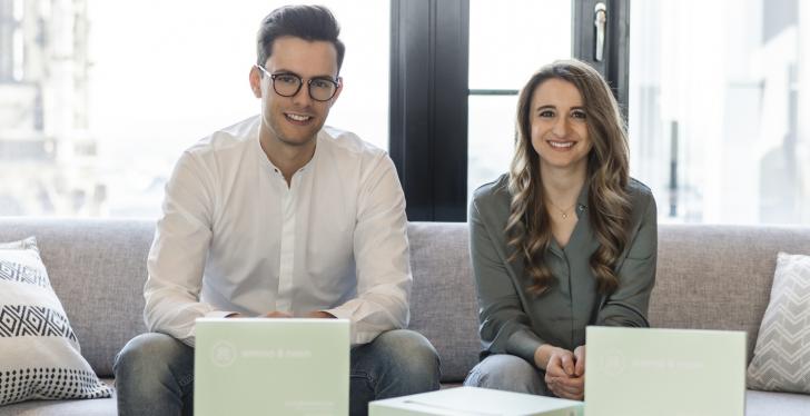 A young man and a young woman sit on a couch in front of a large window and...