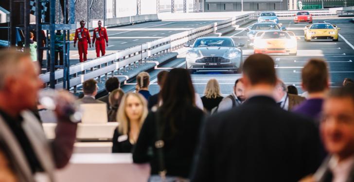 Many visitors at an event, in the background a huge display showing race cars;...