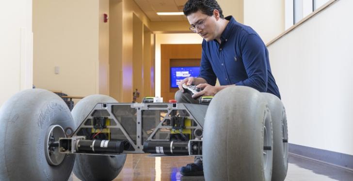 A man is kneeling in a corridor beside a robotic rover with four big wheels;...