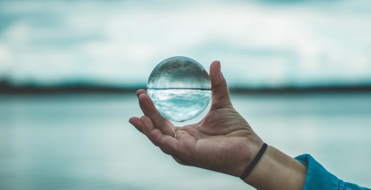 A hand holds a glass ball in which a lake and a sky are reflected...