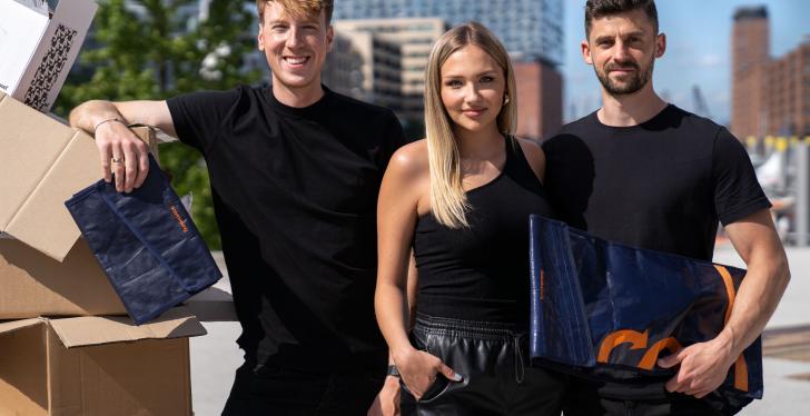 Three people in black clothes stand next to a stack of empty boxes....