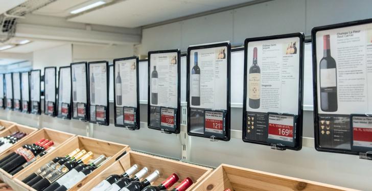 A row of crates with wine bottles, above them electronic price boards...