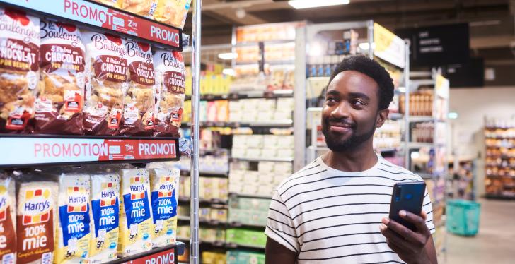 A man with a smartphone in his hand stands next to a supermarket shelf...