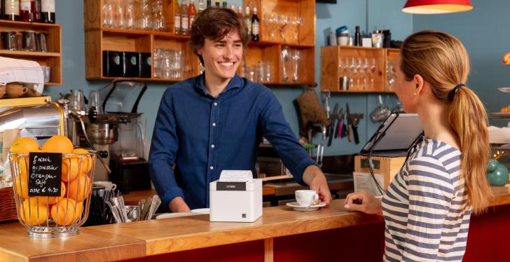 Two people talking at a counter with a POS printer on it...