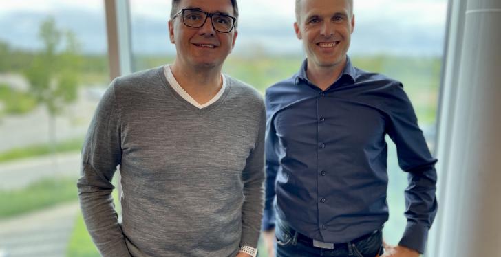 A man with glasses and a grey jumper stands next to a man with a blue shirt,...