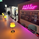 Bar area with yellow table lamps and pink neon lettering reading “Milchbar”....