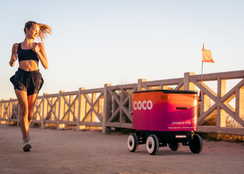 A delivery robot is driving down a walkway beside a female jogger...