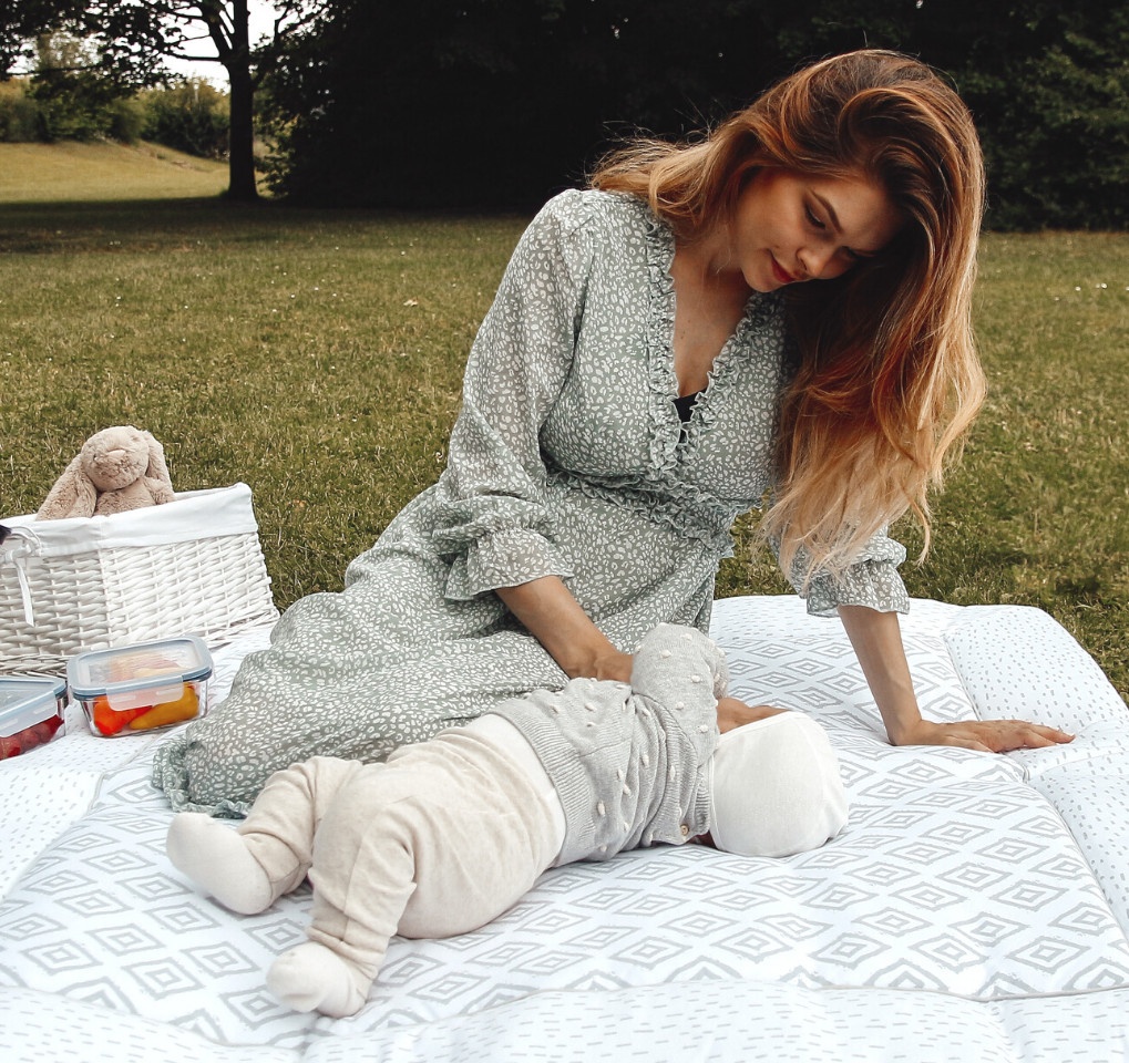 A young mother sits on a picnic blanket in a park, her baby lies in front of her...