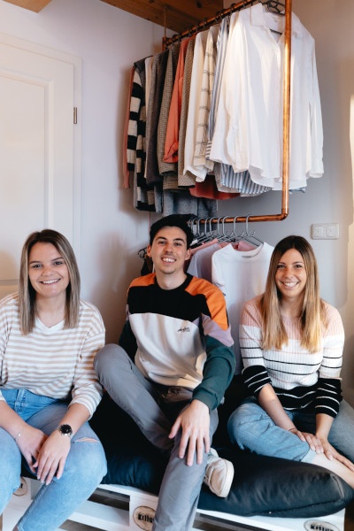 The Retury team sits on a white painted pallet, with tops hanging on a clothes...