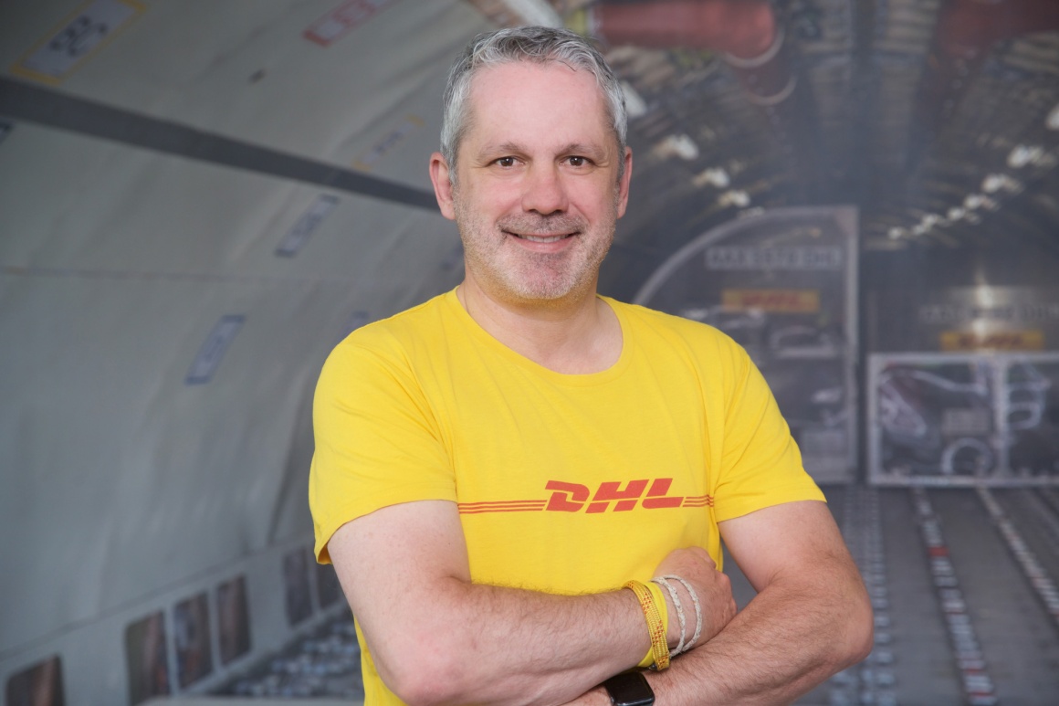 A man with folded arms in a yellow DHL T-shirt in front of the interior of a...