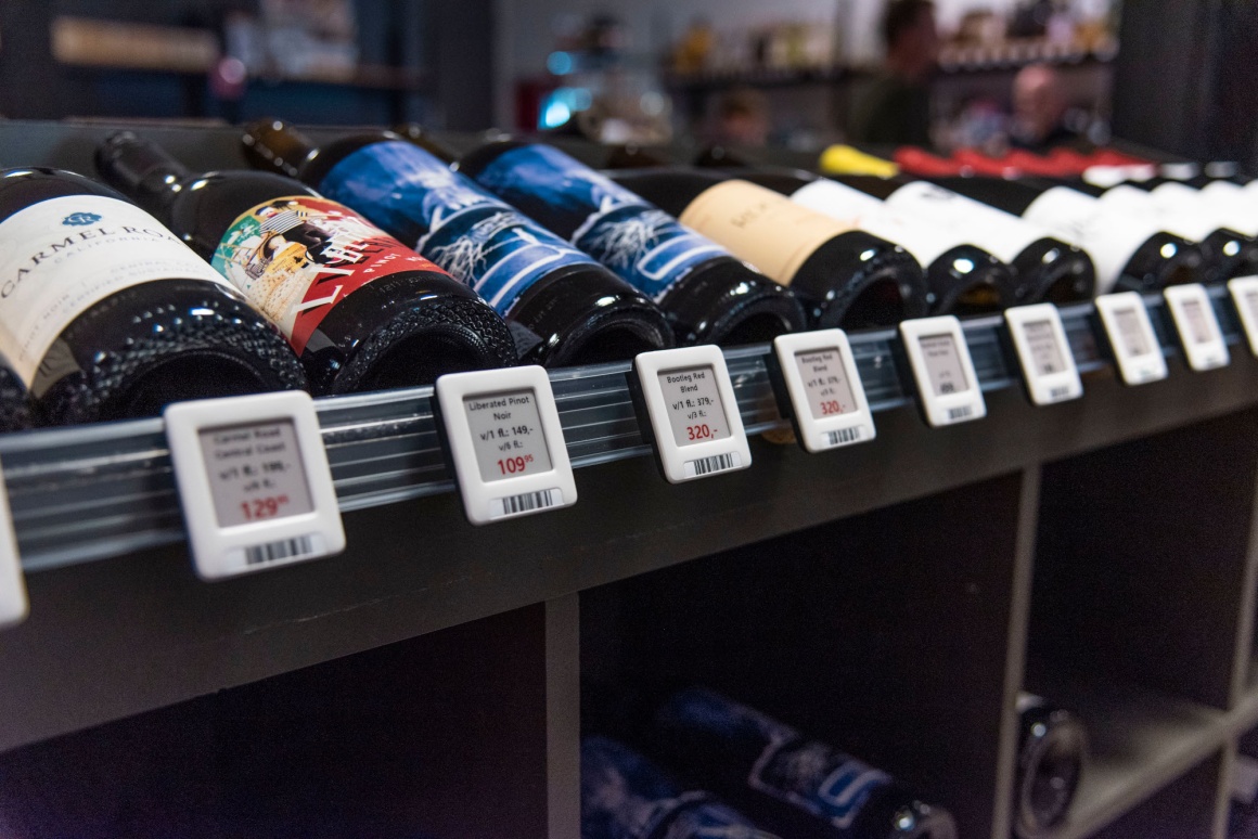 Various wine bottles lie on a shelf. Electronic shelf labels are attached to...