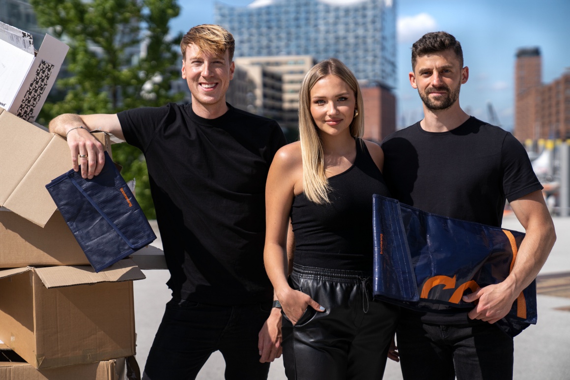 Three people in black clothes stand next to a stack of empty boxes....