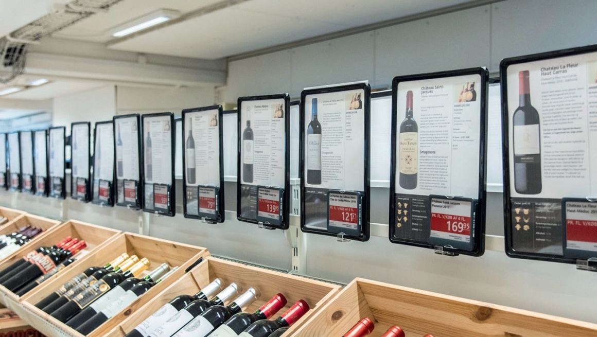 A row of crates with wine bottles, above them electronic price boards...