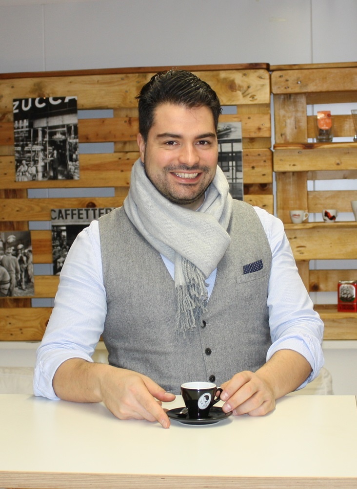 Man in grey waistcoat standing at a table with a coffee cup...