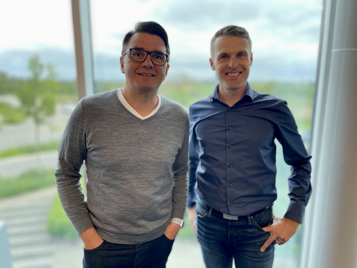 A man with glasses and a grey jumper stands next to a man with a blue shirt,...