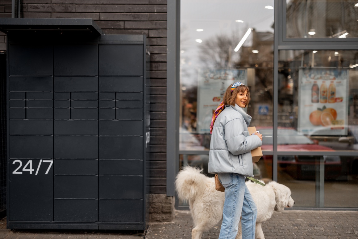 A person with a dog walks past a parcel station to pick up a package....