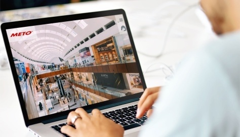 A man is sitting at his laptop. The Meto International GmbH website is open on...