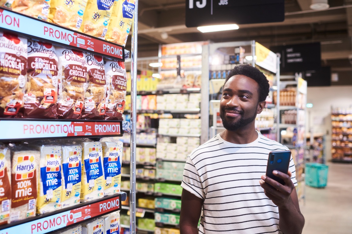 A man with a smartphone in his hand stands next to a supermarket shelf....