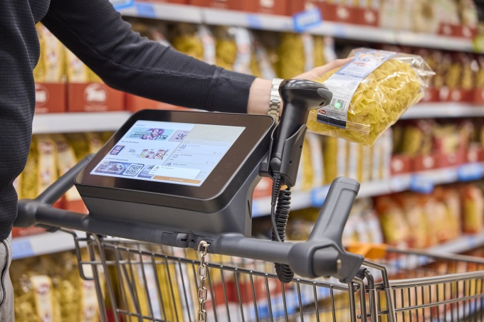 A person holds a packet of pasta in front of the scanner by the shopping trolley...