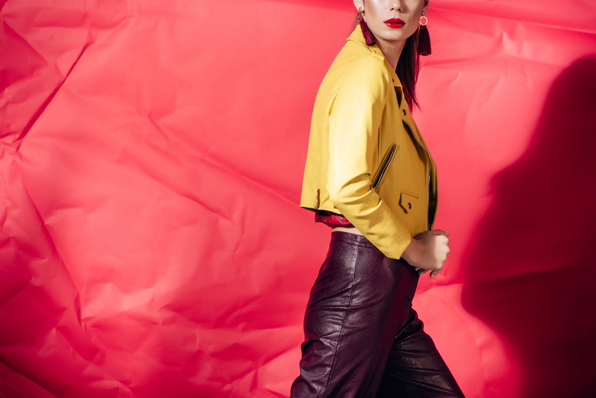 Woman wearing and showcasing a yellow leather jacket in front of a red...