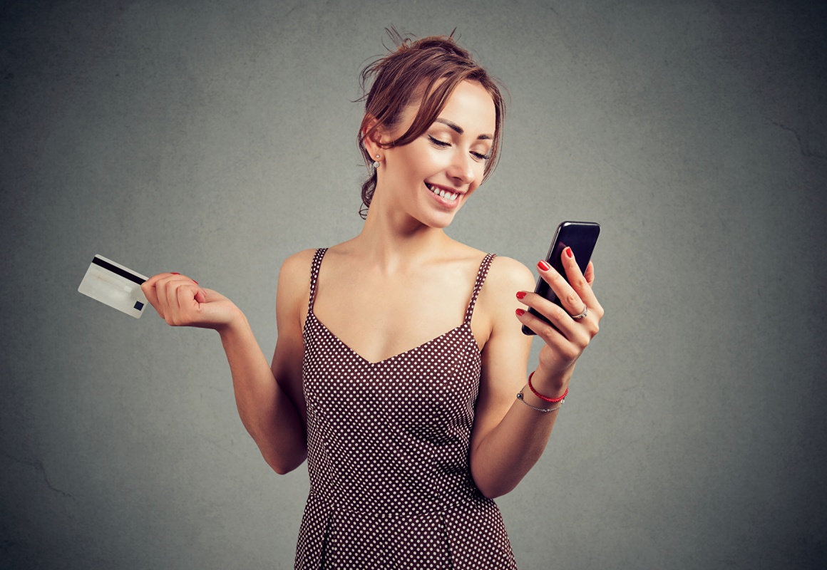 Woman in a dress is smiling at her smartphone and has a card in her hand;...