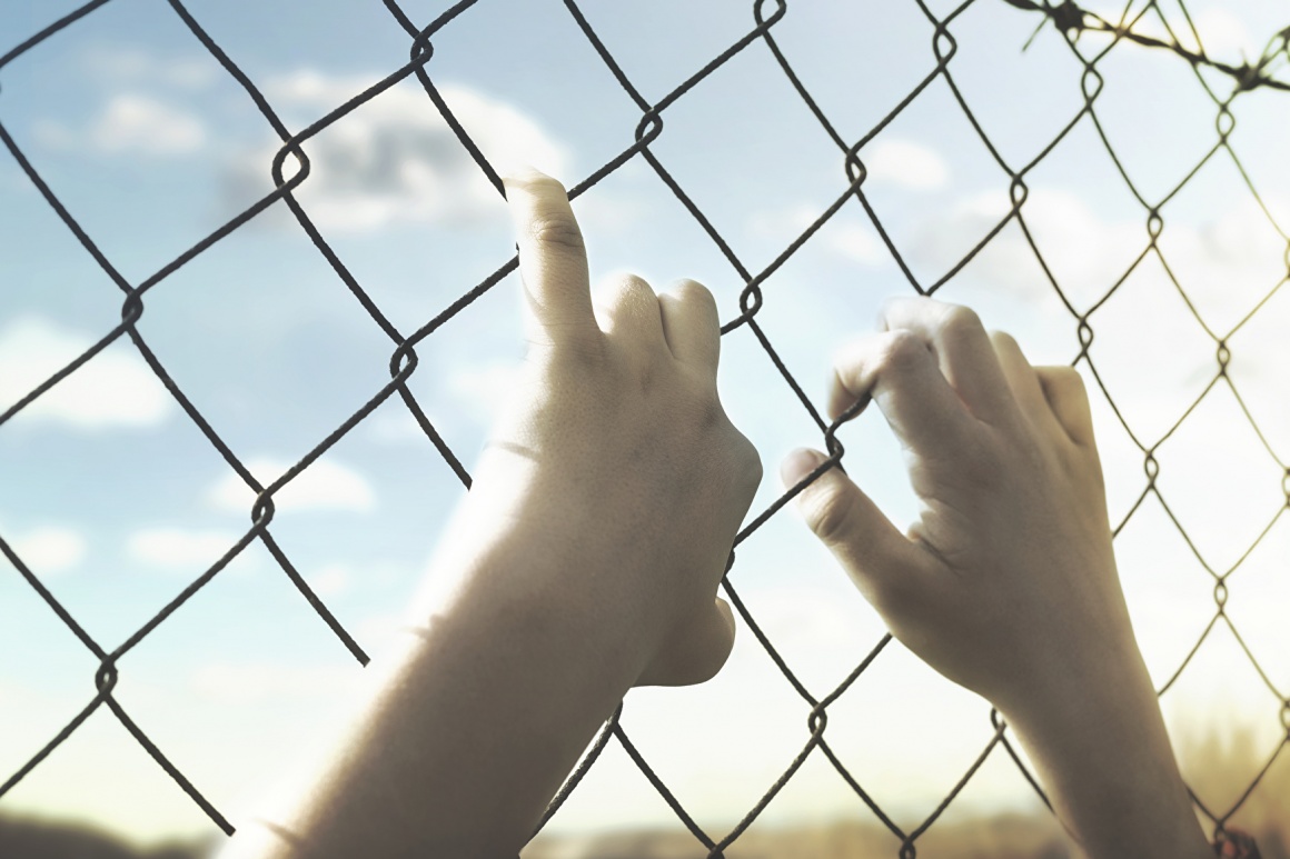 Hands holding onto a wire mesh fence, sunshine behind the fence; copyright:...