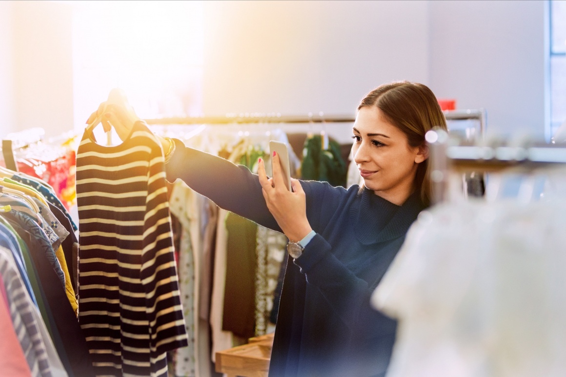 Woman looking at clothing in a store and taking a picture with her smartphone;...