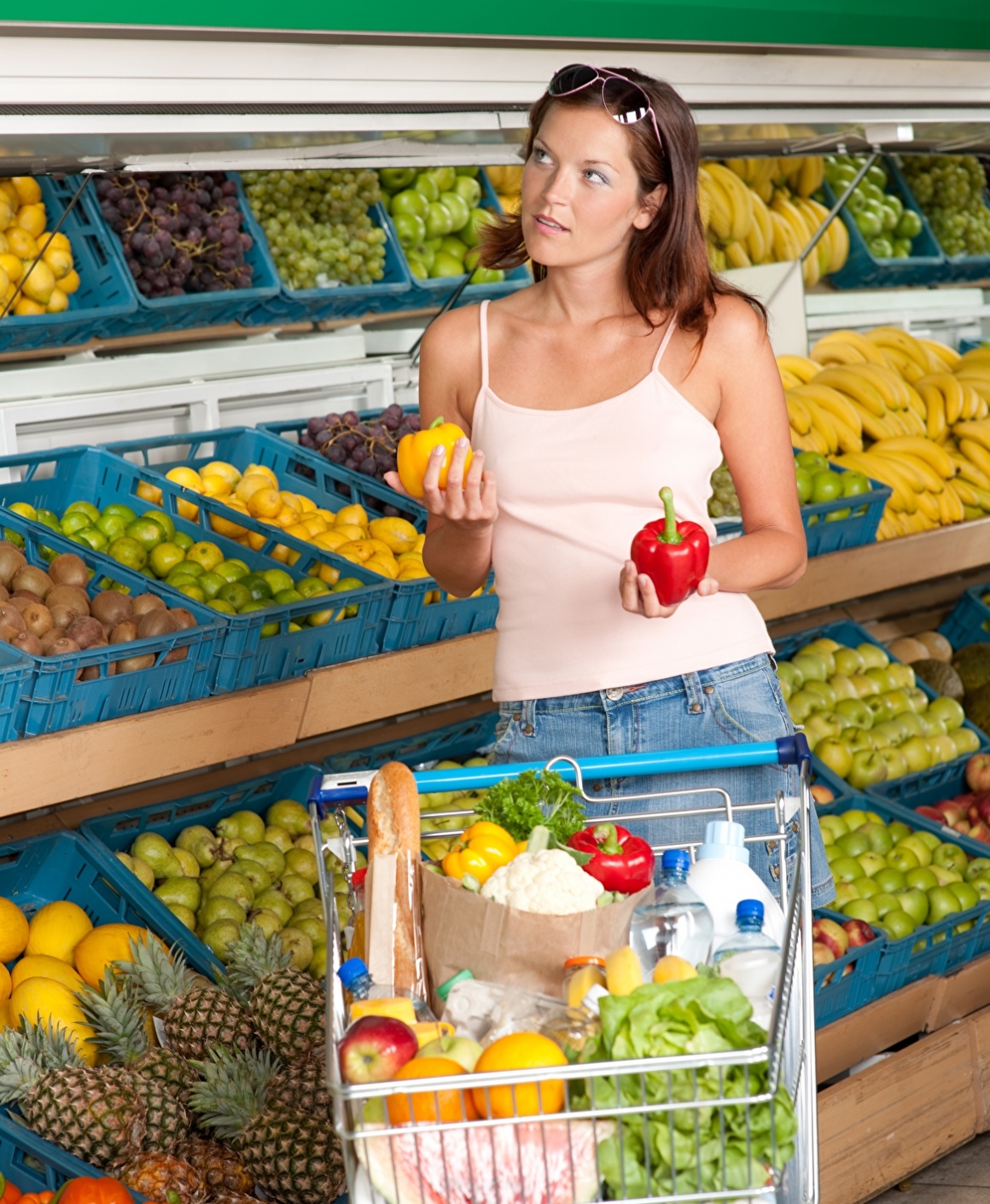 Woman with shopping cart in supermarket holding red and yellow peppers;...