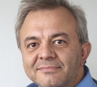 Man with grey hair in light blue sweater smiles into the camera...