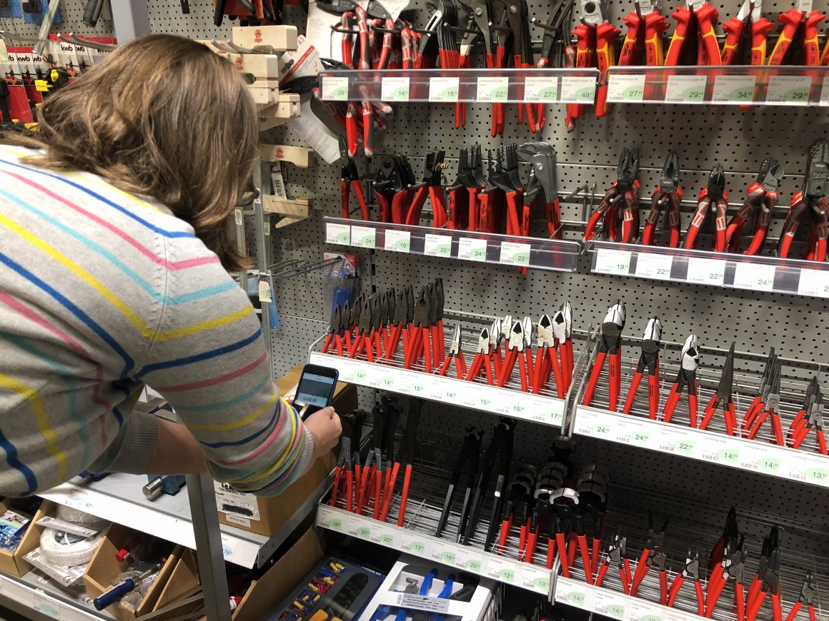 Person holds smartphone to a shelf with pliers and price labels; copyright:...