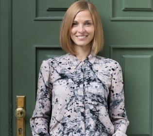 Blonde woman in blouse with flower patterns in front of dark green door smiling...