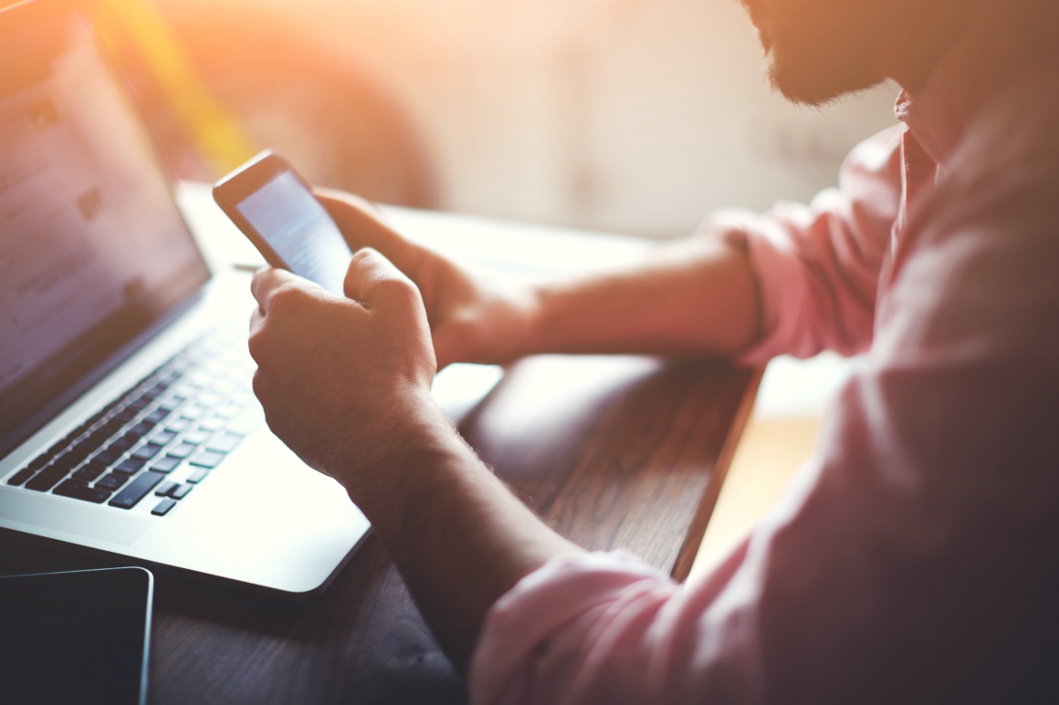 A mans hands hold a smartphone firmly in front of a laptop....