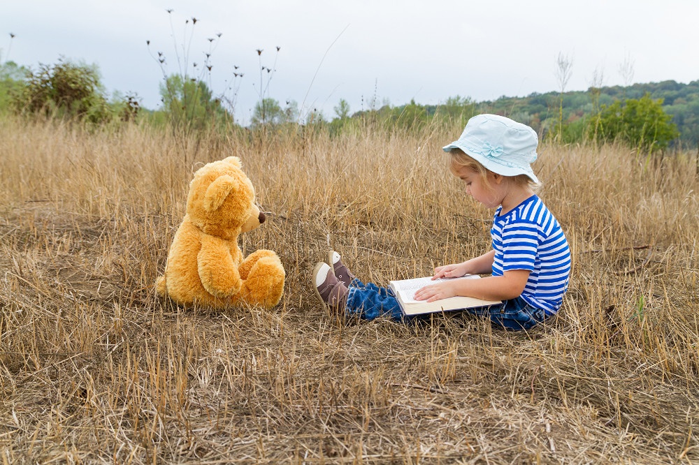 Small child sits with teddy bear and book in the grass and reads aloud...