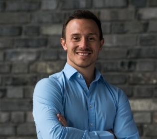 A man in a blue shirt stands and smiles into the camera...