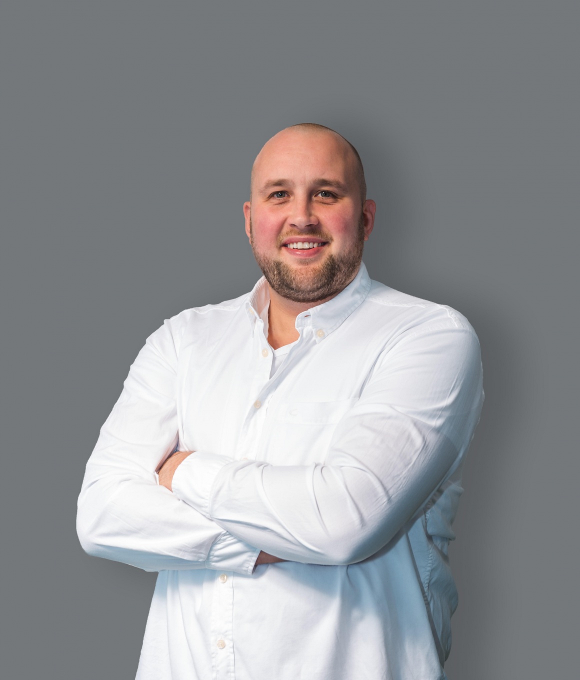 A man in a white shirt in front of a grey background with crossed arms smiles...