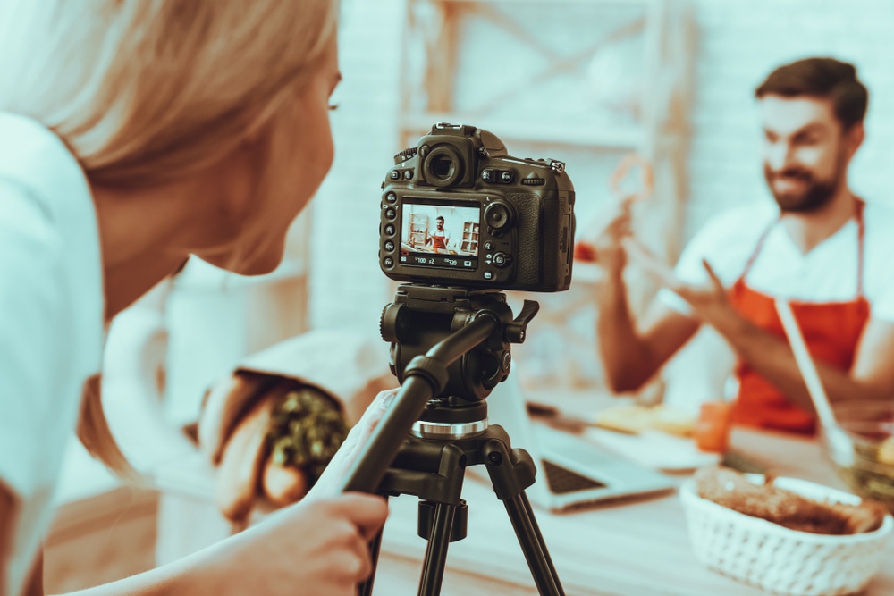 A young blonde woman films with a reflex camera a cooking man in an apron...