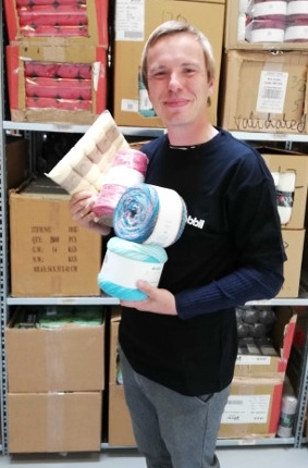 A young man with blond hair holds several packages of wool and smiles into the...